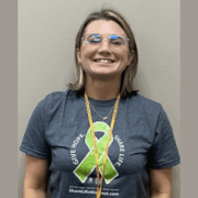 A woman wearing glasses, smiling for a picture, wearing a T-shirt with a green ribbon in support of organ donation.