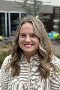 Headshot of Jenna Riley, smiling, wearing a tan and white striped button up blouse