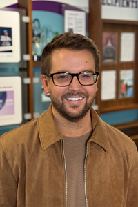 Headshot of John Michael Segars, smiling, wearing a tan shirt and a camel colored jacket